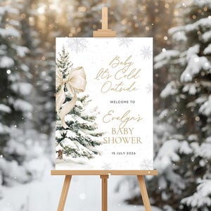 May include: A white sign on a wooden easel with the text "Baby It's Cold Outside" and "Welcome to Evelyn's Baby Shower" in gold script. A watercolor illustration of a snowy pine tree with a bow is on the left.