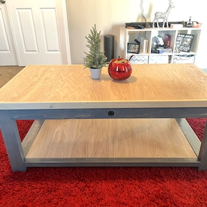 May include: A rectangular coffee table with a light wood top and a gray base. The table has a lower shelf and is decorated with a small Christmas tree and a red decorative ball. The table sits on a red rug.