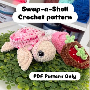 May include: A selection of crocheted toys, including a turtle and a strawberry, are arranged on a bed of green foliage. The image includes the text "Swap-a-Shell Crochet pattern" and "PDF Pattern Only". Colourful yarn is visible in the background.