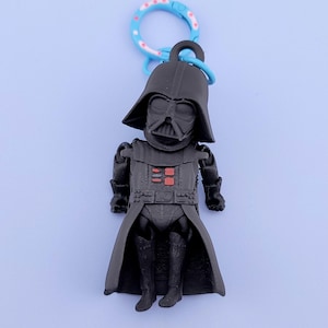 Peut inclure: Un porte-clés noir représentant Dark Vador, avec un fermoir bleu et blanc à motifs. La figurine a un casque noir, une armure de poitrine avec des détails rouges et une longue cape. Un objet de collection Star Wars.