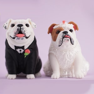 May include: Two bulldog figurines dressed for a wedding. The left bulldog wears a black tuxedo with a black bow tie and a pink rose boutonniere. The right bulldog wears a white lace dress with a matching lace headpiece featuring a small pink heart.