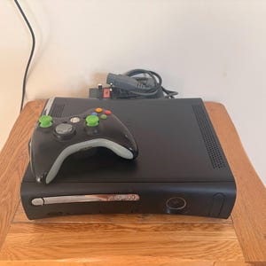 Può includere: Una console per videogiochi Xbox 360 nera con un controller wireless nero abbinato dotato di levette analogiche verde brillante. La console presenta un coperchio argentato per il vano disco con il logo Xbox 360 e un pulsante di accensione.