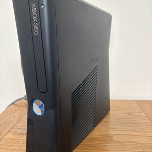 May include: A black Xbox 360 video game console featuring a vertical orientation, a disc drive slot, and a circular power button with a glowing blue light. The side panel includes a ventilation grille and the embossed text Xbox 360.