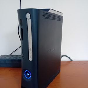 May include: A black Xbox 360 console with a silver trim and the Xbox 360 logo. The console has a blue power button and a matching power supply. The console is on a wooden surface.