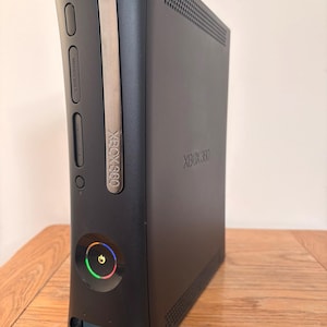 May include: A matte black Xbox 360 video game console standing vertically. The front panel features a silver disc drive cover, two memory unit slots, and a circular power button illuminated with a glowing green ring. The Xbox 360 logo is visible.