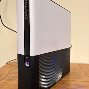 May include: A white and black Xbox 360 video game console standing vertically. The front panel features the Xbox 360 logo in black text, a power button with a blue light, and a glossy white upper section contrasted by a matte black lower section.