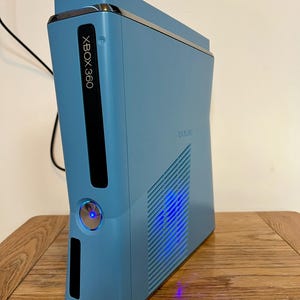 May include: A light blue Xbox 360 video game console featuring a vertical orientation, a black vertical disc drive panel with white text, and a circular power button illuminated with a bright blue light, alongside a side ventilation grille.