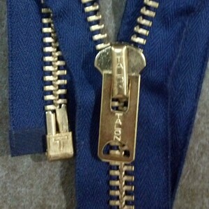 May include: A close-up of a gold zipper with a blue fabric background. The zipper pull has the text "YKK #40Z" on it.