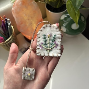May include: A white felt tag and small square, both hand-stitched with a floral design in green, purple, and yellow thread. The tag has a brown string attached. The background includes a lit orange salt lamp and green plants.