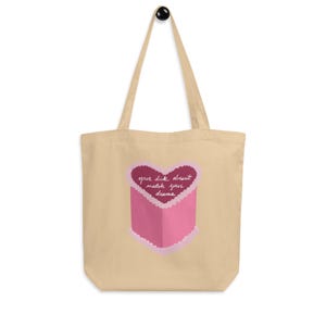 May include: Beige tote bag with a pink heart-shaped design and the text "your dick doesn't match your drama." The design is a pink heart with a pink box. The bag has a black handle.