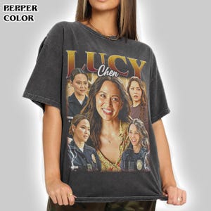 May include: A charcoal gray short-sleeve t-shirt featuring a graphic print of the character Lucy Chen. The design includes the text "LUCY" in large yellow letters and "Chen" in white script, with multiple images of the character.