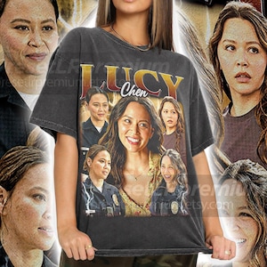 May include: A dark gray t-shirt featuring a collage of images of Lucy Chen, with the name "LUCY Chen" in gold lettering. The design includes multiple images of the actress, creating a vintage, distressed look. The shirt is worn by a person.