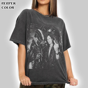 Könnte beinhalten: Ein anthrazitfarbenes Kurzarm-T-Shirt mit einem schwarz-weißen Grafikdruck, der die Alien-Kreatur neben einer Person in einer Lederjacke zeigt, die den Mittelfinger zeigt. Das T-Shirt hat ein verwaschenes Finish im Vintage-Stil.