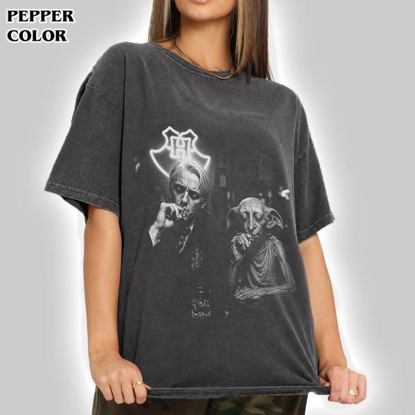 May include: A pepper-colored short-sleeve t-shirt featuring a graphic print of a person and a creature. Above them is a glowing white outline of a crest. The text PEPPER COLOR is printed in the top left corner of the image in black lettering.