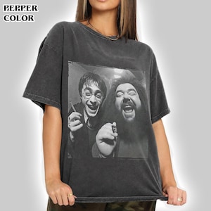 May include: A charcoal gray short-sleeve t-shirt featuring a black and white graphic print of Harry Potter and Rubeus Hagrid laughing. The shirt has a crew neckline and a vintage-style washed finish. The text PEPPER COLOR is printed in the top left corner.