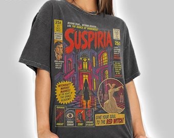 Puede incluir: Una camiseta color pimienta con un gráfico de estilo cómic vintage para la película Suspiria. El diseño incluye ilustraciones en rojo, amarillo y negro de una academia de baile, una figura con vestido blanco y el texto Suspiria.