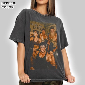 Pode incluir: Uma t-shirt de manga curta em cinzento-carvão com uma impressão gráfica de quatro figuras musculosas num ginásio. O texto "THE BOY WHO LIFTED" está impresso acima das figuras. A camisola tem um decote redondo e um corte largo.