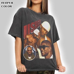 May include: A charcoal grey short-sleeve t-shirt featuring a vintage-style graphic print of the artist Usher. The design includes the text "USHER" in large red letters, multiple photographic portraits of the artist, and a circular inset image.