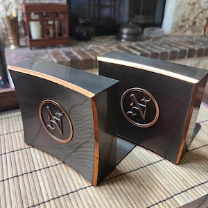 May include: A pair of dark gray bookends with a smooth, polished finish and metallic copper-toned edges. Each bookend features a circular emblem in the center containing a stylized letter N in a decorative font with a copper-toned border.