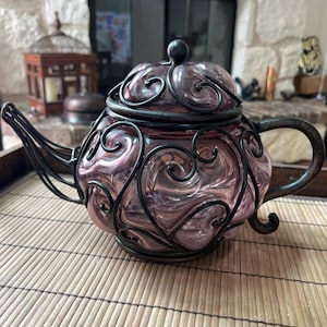 May include: A decorative teapot with a pink glass body and a black metal filigree design. The teapot has a matching lid and handle. The teapot is sitting on a bamboo mat.