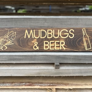 May include: A wooden sign with the text "MUDBUGS & BEER" in gold lettering. The sign also features a drawing of a crawfish and a beer bottle.