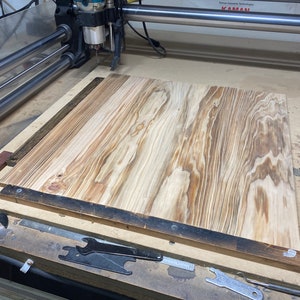 May include: A close-up of a CNC router with a piece of wood on the table. The router is a JET brand and the wood is a light brown color with dark brown streaks. The table is made of metal and has a black finish. The router is in the process of cutting the wood.