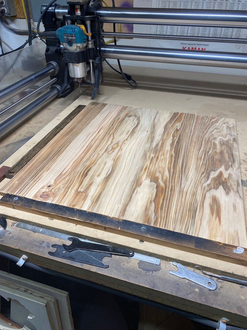 May include: A close-up of a CNC router with a piece of wood on the table. The router is a JET brand and the wood is a light brown color with dark brown streaks. The table is made of metal and has a black finish. The router is in the process of cutting the wood.