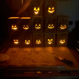 May include: A collection of 12 wooden jack-o'-lantern candle holders with carved faces. The candle holders are stacked in a pyramid shape and lit with warm, glowing lights.