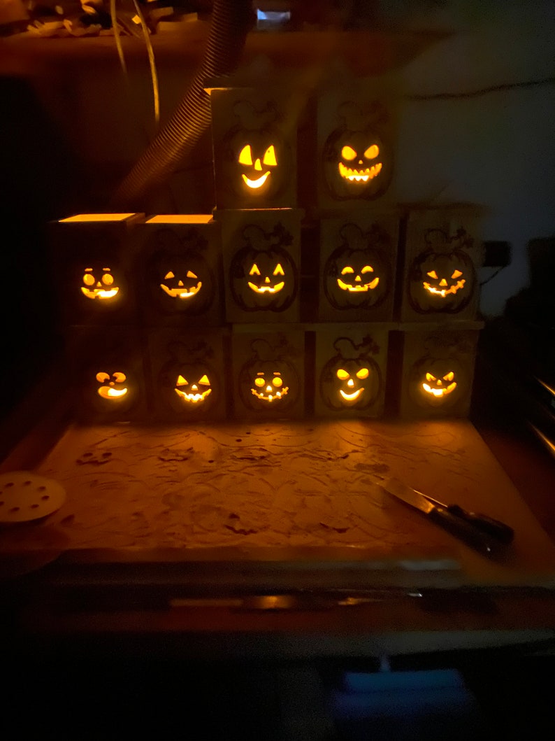 May include: A collection of 12 wooden jack-o'-lantern candle holders with carved faces. The candle holders are stacked in a pyramid shape and lit with warm, glowing lights.
