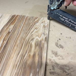 May include: A close-up of a nail gun being used to attach a piece of wood to a larger piece of wood. The nail gun is silver and black with the text "NT 50AE2 21/63" printed on the side.