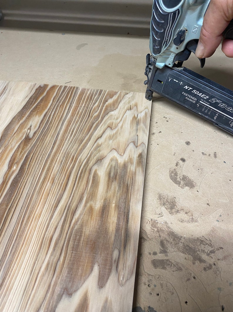 May include: A close-up of a nail gun being used to attach a piece of wood to a larger piece of wood. The nail gun is silver and black with the text "NT 50AE2 21/63" printed on the side.