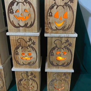 May include: Six wooden pumpkin-shaped boxes with carved jack-o'-lantern faces. The boxes are stacked in two columns of three. Each box has a warm orange glow emanating from the carved face.
