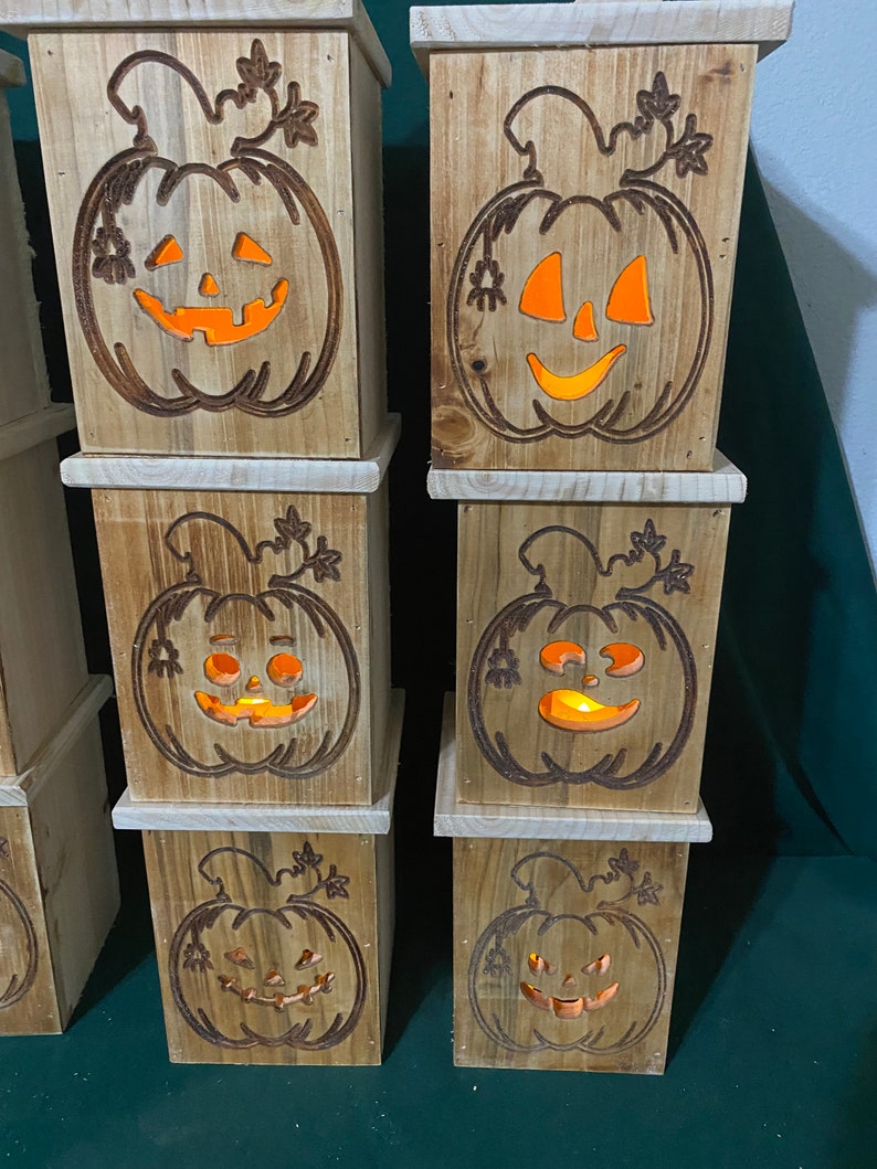 May include: Six wooden pumpkin-shaped boxes with carved jack-o'-lantern faces. The boxes are stacked in two columns of three. Each box has a warm orange glow emanating from the carved face.