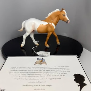Puede incluir: Figura detallada de un caballo con pelaje blanco y marrón, de pie sobre una base circular negra. El caballo tiene crin y cola blancas. Una etiqueta está adherida a la base. Un papel con texto está en primer plano.