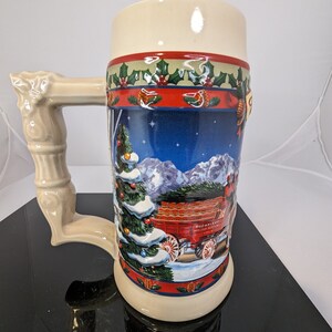 Può includere: Una boccale di birra in ceramica con una scena natalizia che presenta un carro rosso pieno di bottiglie di birra e un paesaggio innevato. La boccale ha un manico bianco e un bordo verde, rosso e bianco.