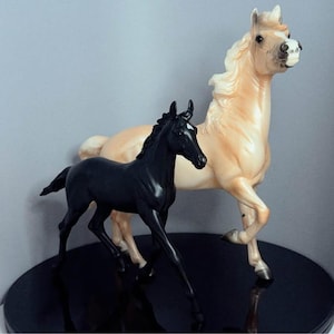 Puede incluir: Dos figuras de caballos. Un potro negro está junto a un caballo adulto de color beige con una melena y cola fluidas. Las figuras están posadas sobre una superficie negra reflectante, con un fondo gris.