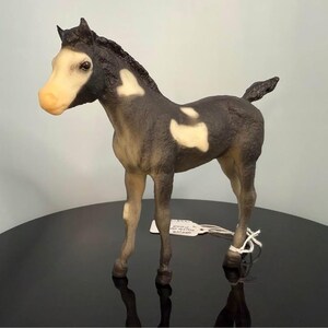 Puede incluir: Figura detallada de un potro, con un cuerpo negro y gris con manchas blancas. El caballo tiene la cara y las patas de color marrón claro. La figura está sobre una superficie negra reflectante.