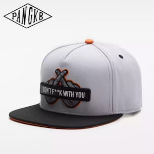 May include: A gray and black baseball cap with an orange brim. The front of the cap has a black patch with the text "I DON'T F**K WITH YOU" and a graphic of two crossed fingers.