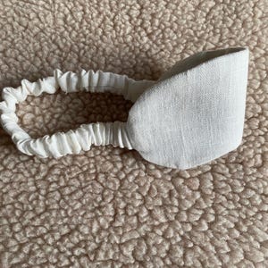 Può includere: Una mascherina per gli occhi in lino bianco con fascia elastica abbinata. La mascherina è progettata per bloccare la luce e favorire il relax. Lo sfondo è una superficie strutturata marrone chiaro.