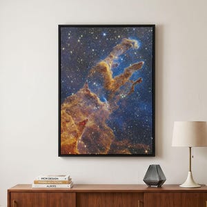 Op de afbeelding: Een ingelijste muurkunstprint met de Pillars of Creation nevel, met levendige oranje, gouden en diepblauwe kosmische stofwolken tegen een achtergrond van verspreide witte sterren, gevat in een dunne zwarte lijst voor presentatie.