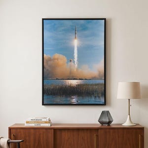 May include: A framed wall art print depicting a rocket launching into a blue sky, leaving a trail of white smoke and orange exhaust clouds above a body of water with tall grasses in the foreground, displayed in a black rectangular frame.