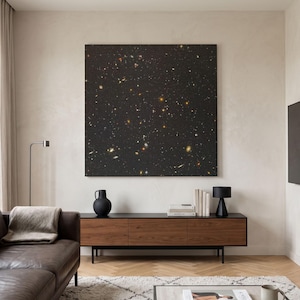 May include: A large square wall art piece featuring a deep black space scene filled with numerous glowing stars and distant galaxies in shades of white, yellow, and orange, mounted centrally on a plain light-coloured wall above a wooden console.