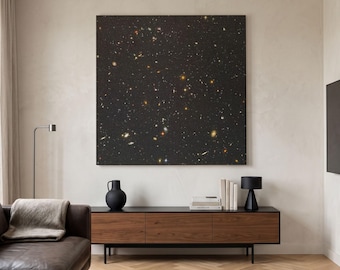 May include: A large square wall art piece featuring a deep black space scene filled with numerous glowing stars and distant galaxies in shades of white, yellow, and orange, mounted centrally on a plain light-colored wall above a wooden console.
