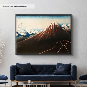 Op de afbeelding: Zwart ingelijste canvas art print met een berglandschap met een besneeuwde top. Het kunstwerk toont een berg met een donkerbruine basis en een witte top, tegen een blauwe lucht met witte wolken. De print is ingelijst in een zwarte rand.