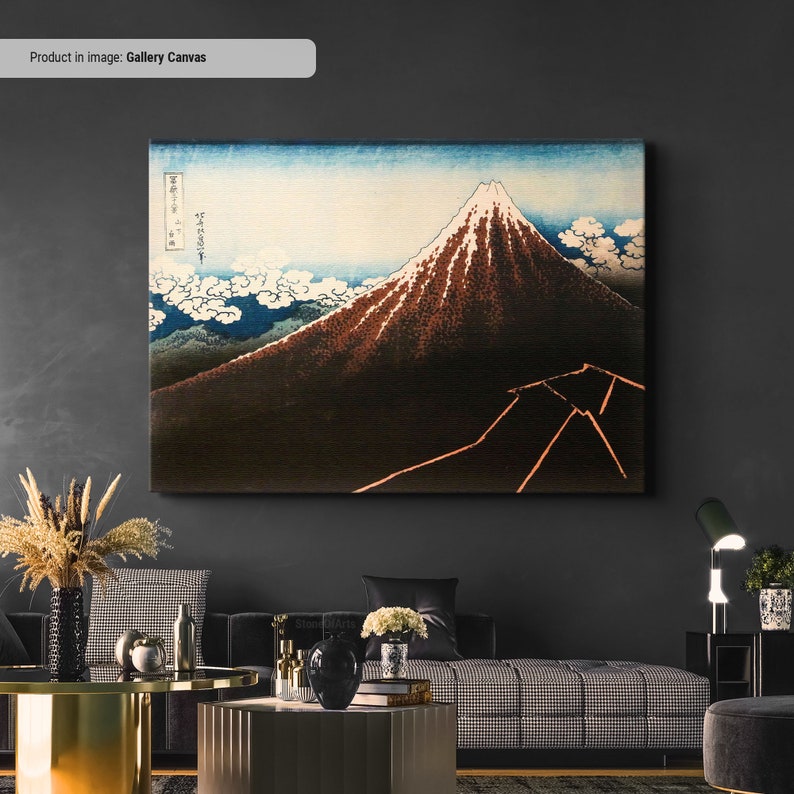 Op de afbeelding: Een ingelijste canvas print van een Japans landschap met de berg Fuji. Het kunstwerk toont de berg met een besneeuwde top, tegen een blauwe lucht met witte wolken. Het canvas is rechthoekig en aan een donkere muur bevestigd.
