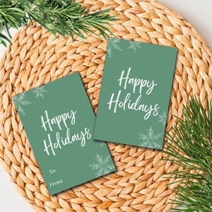 Puede incluir: Dos etiquetas de regalo verdes con copos de nieve blancos y el texto "Felices Fiestas". Una etiqueta tiene espacio para "Para:" y "De:"