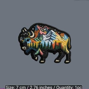 May include: An embroidered patch shaped like a bison, featuring a landscape scene with mountains, pine trees, and a night sky with stars and a crescent moon. The patch measures 2.76 inches in width and is rendered in black, blue, green, and yellow.