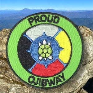 Può includere: Toppa rotonda con la scritta "PROUD OJIBWAY". Il design centrale presenta una tartaruga circondata da segmenti blu, gialli, rossi, neri e bianchi. Bordo verde. Sullo sfondo, una catena montuosa.