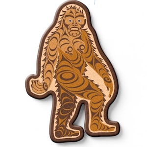 Può includere: Un oggetto decorativo a forma di Bigfoot marrone e beige con un design tribale dettagliato. La figura ha un bordo marrone scuro e presenta un viso e un corpo stilizzati con motivi intricati. Il design ricorda l'arte nativa americana.