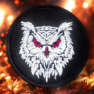 May include: Embroidered patch with an owl design. The owl's face is white against a black circular background. The owl has red eyes and a red beak. The patch is a circular shape, suitable for sewing onto clothing or bags.